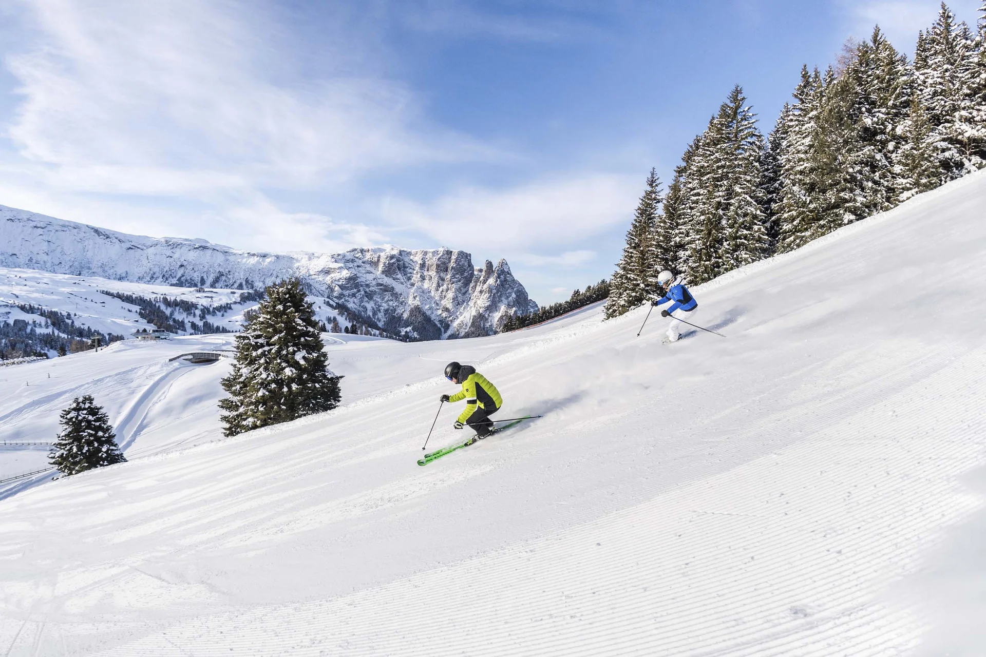 Genuss und Wellness à la Martina Zwei Skifahrer fahren einen schneebedeckten Hang mit Tannen im Hintergrund hinunter
